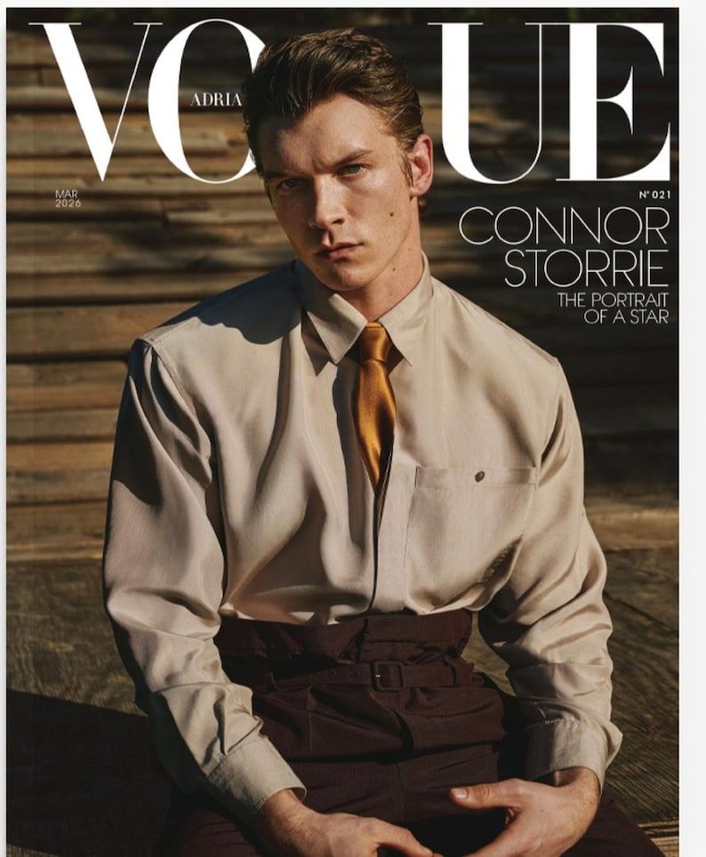 May include: Vogue magazine cover featuring Connor Storrie. The cover has the Vogue logo in white, with the text "Adria" above the image. The model is wearing a beige button-down shirt with a gold tie and brown pants. The text "The Portrait of a Star" is also visible.