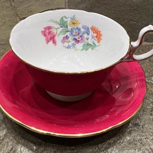 Puede incluir: Un juego de taza y platillo de té vintage. La taza y el platillo son de un rosa fucsia vibrante con ribete dorado. El interior de la taza blanca presenta un ramo floral de flores rosas, amarillas, azules y naranjas. El asa es blanca con detalles dorados.