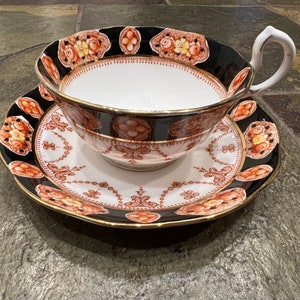 Puede incluir: Un juego de taza y platillo de té negro y naranja con un diseño floral. La taza tiene un interior blanco y un borde dorado. El platillo tiene un borde festoneado.