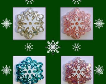 May include: Four snowflake-shaped cover minders in gold, pink, teal, and purple glitter. Each has a white center and is set against a green background with the text "SNOWFLAKE COVER MINDERS". The logo "TRACY'S - DP WORLD" is in the bottom right.