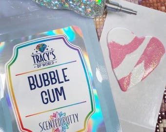 May include: A light blue pouch of bubble gum scented putty with a holographic label reading TRACY'S DP WORLD BUBBLE GUM SCENTED PUTTY. Next to it is a heart-shaped piece of pink and white glittery putty and a silver glittery diamond painting pen.
