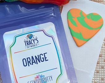 May include: A purple pouch of orange scented putty with a label reading TRACY'S DP WORLD ORANGE SCENTED PUTTY. Next to the pouch is a heart-shaped piece of orange and green putty and a multi-colored tool with a metal tip. The label features a diamond icon.