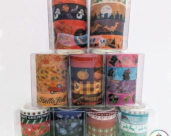 May include: A collection of holiday washi tape sets stacked in clear plastic boxes. The tapes feature seasonal designs including Halloween ghosts, pumpkins, Thanksgiving motifs, autumn leaves, Christmas reindeer, snowflakes, and festive patterns.