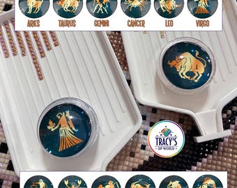 May include: A set of round zodiac-themed covers for diamond painting trays, featuring gold-colored astrological symbols on a dark blue starry background. The text LILYPADS - ZODIACS, CHOOSE YOUR SIGN - INCLUDES BLACK AND ORANGE SPONGES, and TRACY'S DP WORLD are visible.