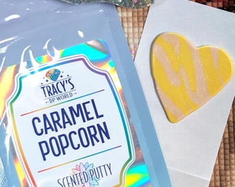 May include: A light blue pouch with a holographic label reading TRACY'S DP WORLD CARAMEL POPCORN SCENTED PUTTY, accompanied by a heart-shaped piece of yellow and beige swirled putty and a glittery tool with a metal tip on a white card.