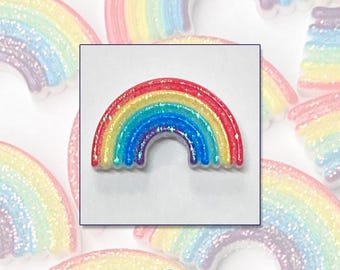 May include: A colorful rainbow-shaped cover minder with glitter accents. The cover minder is in a square frame, with other rainbow shapes in the background. The text "RAINBOW COVER MINDER" is at the top of the image.