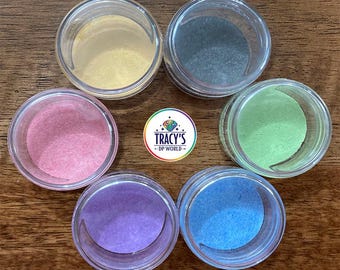 May include: Six small, clear jars filled with colorful powders, arranged in a circle. The powders are in shades of yellow, gray, pink, green, purple, and blue. Text at the top reads "LARGE LILYPADS" and at the bottom "FITS LARGER MUTI-PLACERS - CHOICE OF 6 COLORS".