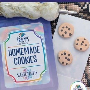 May include: A purple pouch labeled HOMEMADE COOKIES SCENTED PUTTY with a rainbow border, featuring a white and gold marbled stylus tool at the top and three small tan circular clay pieces with black dots resembling cookies on a white card.