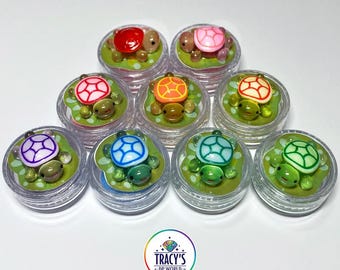 May include: A collection of small, clear containers each holding a miniature turtle figurine on a lilypad. The turtles have colorful shells in shades of red, pink, orange, yellow, purple, blue, and green. The text "LILYPADS - TURTLES" is at the top.