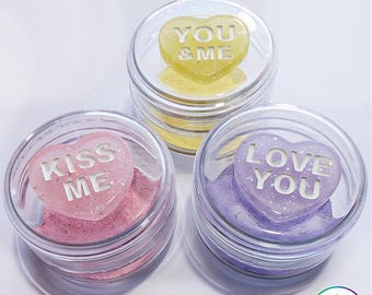 May include: Three clear circular containers, each topped with a glittery heart-shaped lid. The lids feature the phrases YOU & ME in yellow, KISS ME in pink, and LOVE YOU in purple, with matching colored sponges visible inside each container.