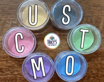 May include: Six round clear containers filled with colored powder, arranged in a circle. The containers hold gold, silver, pink, green, purple, and blue powder. Large white letters U, S, T, C, M, O are overlaid on the containers. A logo for Tracy's DP World is in the center. Text reads CUSTOM LARGE LILYPADS and FITS LARGER MULTI-PLACERS - CHOICE OF 6 COLORS.