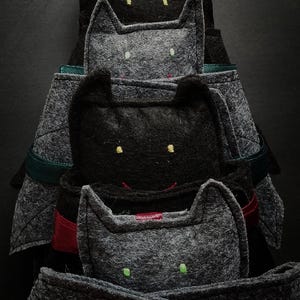 May include: A stack of handmade cat-shaped felt ornaments in varying shades of grey and black. Each cat has embroidered eyes and a small smile. Some cats have red or green ribbon accents.