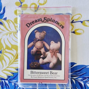 Könnte beinhalten: Eine rosa Verpackung mit der Aufschrift "Dream Spinners" und "Bittersweet Bear". Die Packung enthält ein Schnittmuster für einen 76 cm großen Bären mit Quilt- und Tee-Färbeanweisungen. Das Bild zeigt zwei Bärenillustrationen, eine mit einer Schleife und die andere mit einem Muster.