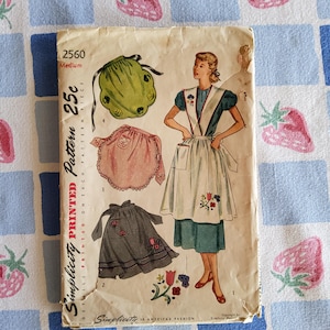 May include: Vintage Simplicity pattern envelope featuring apron designs. The cover shows illustrations of aprons in green, pink, and gray, with floral embellishments. The text on the envelope reads "Simplicity Printed Pattern 25c".
