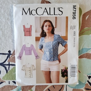 May include: A McCall's sewing pattern envelope, M7956, featuring illustrations of women's tops. The main image shows a woman wearing a blue top with star patterns and a square neckline. The envelope includes size information: AX5 (4-6-8-10-12).