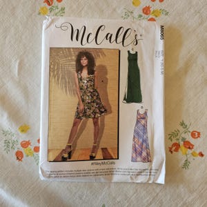Könnte beinhalten: Ein McCall's M8065 Schnittmusterumschlag mit einem ärmellosen Kleid mit tropischem Blumenmuster, einem einfarbigen dunkelgrünen bodenlangen Kleid und einem karierten bodenlangen Kleid. Der weiße Papierumschlag enthält Text und Abbildungen.