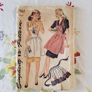 May include: Vintage Simplicity sewing pattern 1162 for a small size apron. The pattern features two women wearing different styles of aprons, one with a floral print and the other with a gingham pattern. The text "Simplicity 1162 Cut to Exact Size Pattern" is printed on the side of the pattern.