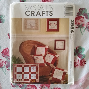 Op de afbeelding: Een McCall's Crafts-patroon voor een quilt met een rood-wit geruite achtergrond en negen verschillende geborduurde vierkanten. De vierkanten bevatten ontwerpen zoals een huis, een hart, een boom en een vogel. Het patroon bevat ook instructies voor het maken van kussens met geborduurde ontwerpen zoals "Home Sweet Home", "Friends warm the heart" en "I believe in Angels".