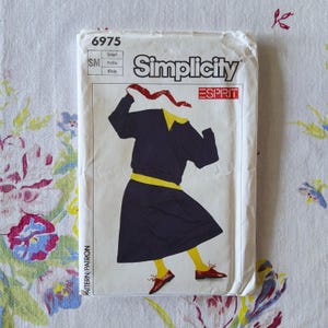 May include: A Simplicity 6975 sewing pattern envelope for a size small petite garment. The illustration shows a dark navy dress with a yellow collar and waistband, yellow tights, and brown shoes, with a red scarf held above the neckline.