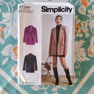 Op de afbeelding: Simplicity naaipatroon R11941 met een double-breasted blazer. Het patroon bevat illustraties van een paarse en een zwarte blazer. Het model op de omslag draagt een roze geruite blazer met zwarte laarzen.