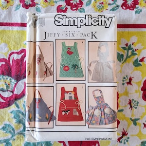 May include: A Simplicity Jiffy Six Pack sewing pattern for aprons. The package features six apron designs, including aprons with chili peppers, a flamingo, a farm scene, and seashells. The package is white with the Simplicity logo and the words "Jiffy Six Pack".