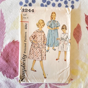 May include: Vintage Simplicity sewing pattern for a girl's robe and dress, size 8. The pattern features illustrations of three different robe and dress designs. The pattern is printed on paper and has a cream-colored background.