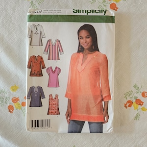 Puede incluir: Un sobre de patrón de costura Simplicity, número 559, con ilustraciones de seis diseños de túnicas diferentes. El frente muestra a un modelo con una túnica naranja de manga tres cuartos con cuello en V y un código de barras en la parte inferior.