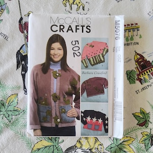 May include: A McCall's Crafts sewing pattern envelope M5076 for a cardigan. The front shows a mauve cardigan with green leaf appliqués and denim pockets, alongside smaller images of a watermelon-themed sweater and a black star-patterned cardigan.