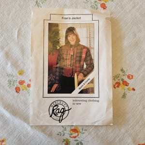 May include: A white sewing pattern envelope titled Fran's Jacket featuring a photograph of a person wearing a red, black, and white plaid jacket. The envelope includes the text Merchant and Mills Rag interesting clothing to sew and Quick & Comfy.