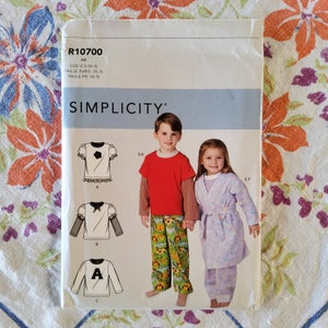 Pode incluir: Um padrão de costura Simplicity para roupas infantis, com uma menina em um roupão roxo e um menino em uma camisa vermelha e calças com estampa de girassol. O número do padrão é R10700 e o tamanho é AA (6-3).