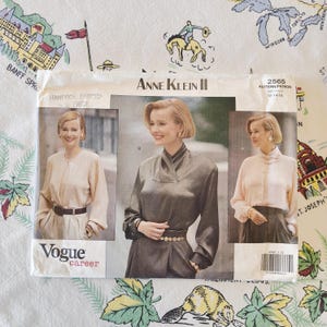 Puede incluir: Un sobre de patrón de costura Vogue Career de Anne Klein II, número 2565, para tallas 12-14-16. El frente muestra tres imágenes de una blusa de manga larga con cuello alto, en rosa claro y gris oscuro, con un código de barras y texto.