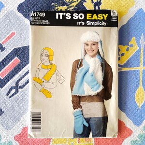 May include: A sewing pattern titled "IT'S SO EASY" by "It's Simplicity". The pattern features a woman wearing a blue and white winter hat, scarf, and mittens. The pattern also includes a drawing of a woman in a yellow hat and scarf.