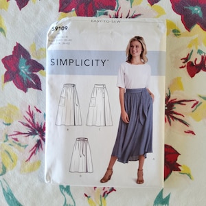 May include: A Simplicity sewing pattern package, featuring a woman in a white top and blue skirt. The package includes illustrations of skirt designs and the text "EASY-TO-SEW" and "SIMPLICITY". The pattern size is US 6-14.