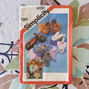 Könnte beinhalten: Simplicity-Schnittmuster 6231 mit verschiedenen Tier-Hausschuhen. Das Cover zeigt Hausschuhe in Form von Löwen, Bären, Kaninchen und Mäusen. Das Muster enthält ein Foto eines Kindes, das die Hausschuhe trägt.