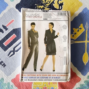 以下が含まれることがあります： グレーのスーツとドレスを着た女性のイラストが描かれた、Burda Internationalの裁縫パターンパッケージ。 パッケージには「KOMBINATION」と「COORDONNEES」の文字が含まれています。 パッケージはカラフルな模様の生地の上にあります。