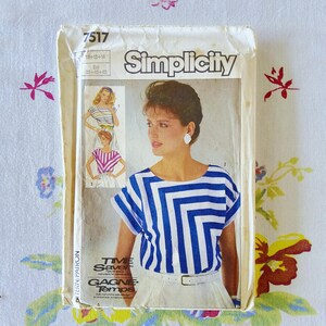 May include: A Simplicity 7517 sewing pattern envelope featuring a white top with blue horizontal and vertical stripes. The cover displays a photograph of a person wearing the top, along with illustrations of two variations, and the text Time Saver.