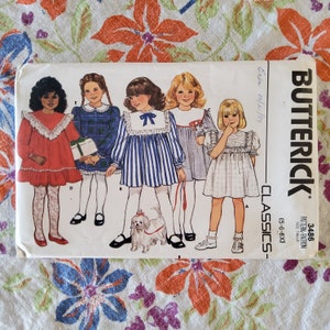 Puede incluir: Un patrón de costura vintage para vestidos de niñas. El patrón es para las tallas 5-6-6X e incluye cinco estilos de vestidos diferentes. Los vestidos son todos de diferentes colores y patrones, incluyendo rojo, azul, blanco y cuadros. El patrón es de Butterick y está fechado el 08/21/84.