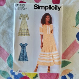 May include: Simplicity sewing pattern R11453 featuring three dress designs. The pattern includes sizes 8-20. The main image shows a woman wearing a yellow dress with white lace trim. The pattern's text includes "Simplicity" and size information.