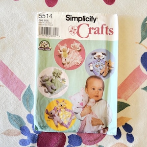 May include: A Simplicity Crafts sewing pattern featuring various baby toy designs. The pattern includes images of stuffed animals, such as a cat, dog, and bear, along with a baby holding a stuffed animal. The pattern's cover is white and green.