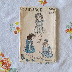May include: Vintage sewing pattern titled "Advance 3410" featuring illustrations of young girls in dresses. The pattern cover is aged with visible wear and tear, and the illustrations depict the dresses with ruffles and bows. The text on the cover includes "Size 2" and "Breast 21".