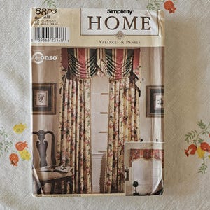 Może przedstawiać: Koperta z wykrojem krawieckim Simplicity Home na lambrekiny i panele, numer 8803. Na froncie znajduje się zdjęcie zasłon w kwiaty sięgających podłogi z pasiastym, drapowanym lambrekinem oraz mniejszy obrazek przedstawiający podobną dekorację.