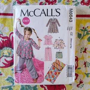 Może przedstawiać: Koperta z wykrojem krawieckim McCall's M6643 dla odzieży w rozmiarze CZ (Med-Lrg-Xlg). Na froncie znajduje się zdjęcie dziecka w kwiecistym topie i wzorzystych spodniach oraz ilustracje różnych stylów odzieży, w tym sukienek i piżam.