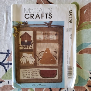 Könnte beinhalten: Ein McCall's Crafts-Muster für eine Garten-Wandbehänge. Das Muster beinhaltet ein Vogelhaus, einen Bienenstock, ein Paar Gartenhandschuhe und eine Wassermelone. Das Muster trägt den Titel "Gärtnern ist Therapie für die Seele".