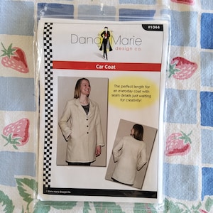 Könnte beinhalten: Ein Schnittmuster für einen Automantel, mit einem weißen Mantel mit Knopfleiste und Kragen. Das Schnittmuster stammt von Dana Marie Design Co. und trägt die Nummer #1044. Der Text lautet "Die perfekte Länge für einen Alltagsmantel mit Nahtdetails, die nur darauf warten, kreativ gestaltet zu werden!"