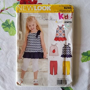 May include: A sewing pattern for kids' clothing, featuring a blue and white chevron print top with a ruffle, a red and white polka dot top, and a yellow pair of pants. The pattern is from New Look and is called "Just 4 Knits".