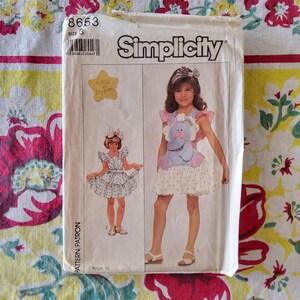 May include: Simplicity sewing pattern for a child's dress with an elephant applique. The pattern envelope is white with illustrations of the dress and the text "Simplicity". The size is 3. The envelope has a star-shaped sticker that says "made in heaven".