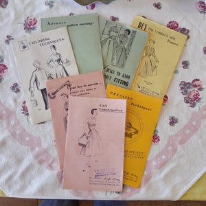 Könnte beinhalten: Eine Sammlung von Vintage-Schnittmusterheften mit Titeln wie "Tailoring Techniques" und "Unit Construction". Die Umschläge zeigen Illustrationen von Kleidung und Texte wie "Buy the Correct Size Pattern". Die Hefte sind auf einer Tischdecke mit Blumenmuster angeordnet.