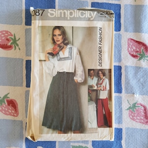 May include: A vintage Simplicity sewing pattern, number 087, for a designer fashion skirt and blouse. The pattern features a woman wearing a white blouse with a navy blue collar and a dark green skirt. The pattern also shows a woman wearing a white blouse with a navy blue collar and red pants.
