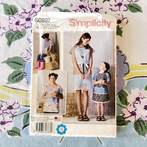 May include: A Simplicity sewing pattern package featuring images of women and children wearing dresses and tops. The package is white with pink and red text and includes the pattern number S0897.