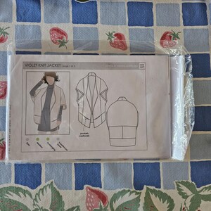 May include: A sewing pattern envelope for a Violet Knit Jacket, enclosed in clear plastic. The front displays a technical illustration of the jacket, including front and back views, optional pocket details, and a difficulty scale labeled Easy to Experienced.